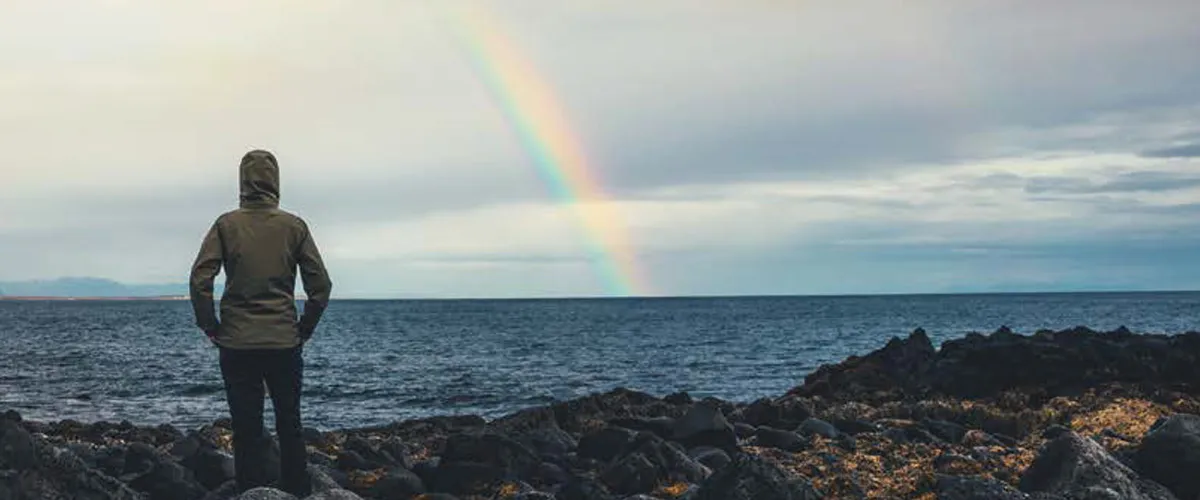 A woman looks out to sea as a rainbow beams from the sky