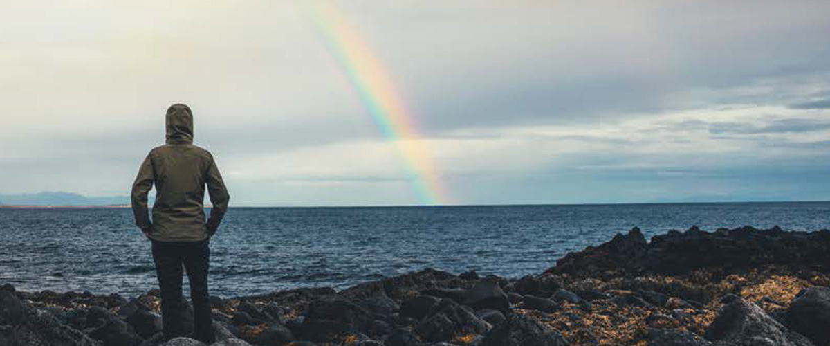 A woman looks out to sea as a rainbow beams from the sky